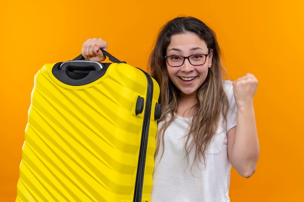 Mujer joven viajero en camiseta blanca con maleta emocionado y feliz apretando el puño de pie sobre la pared naranja