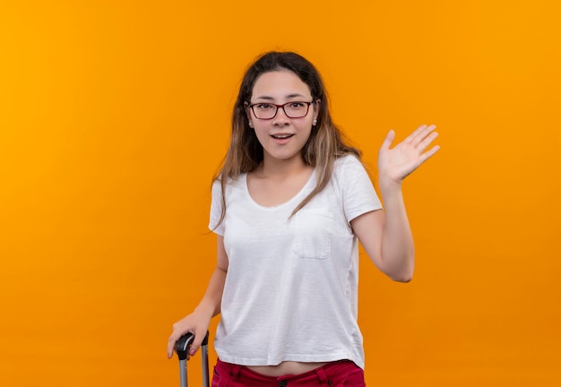 Mujer joven viajero en camiseta blanca con maleta sorprendida y feliz saludando con la mano de pie sobre la pared naranja