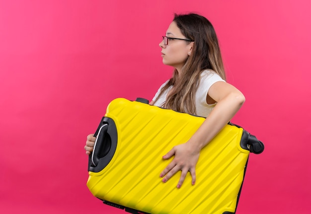 Foto gratuita mujer joven viajero en camiseta blanca con maleta de viaje de pie de lado sobre pared rosa