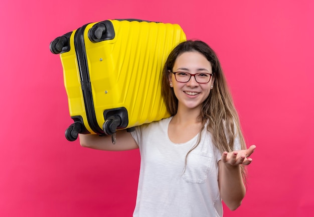Mujer joven viajero en camiseta blanca con maleta de viaje sonriendo alegremente levantando la mano de pie sobre la pared rosa