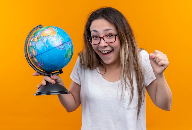 Mujer joven viajero en camiseta blanca sosteniendo globo mirando salido y feliz puño de pie sobre la pared naranja