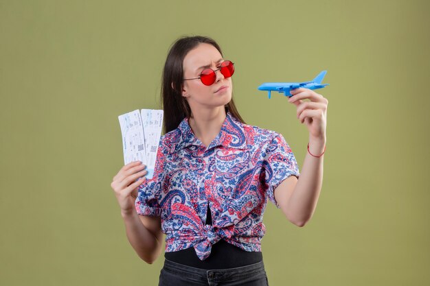 Mujer joven viajero con gafas de sol rojas con boletos y avión de juguete mirándolo con expresión pensativa con el ceño fruncido sobre pared verde