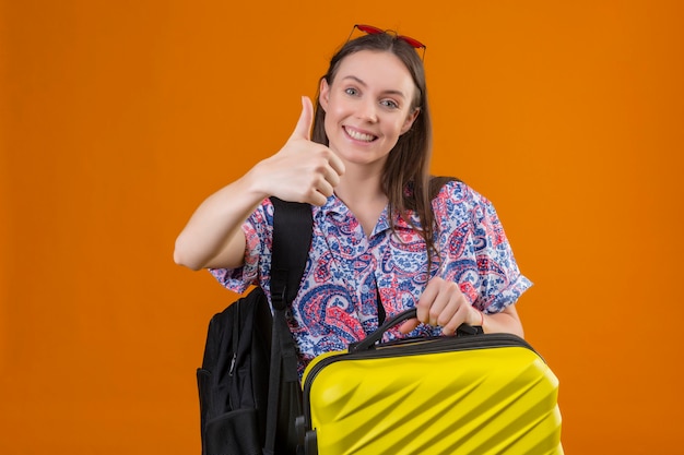 Mujer joven viajero con gafas de sol rojas en la cabeza con maleta con mochila sonriendo alegremente mostrando los pulgares sobre la pared naranja