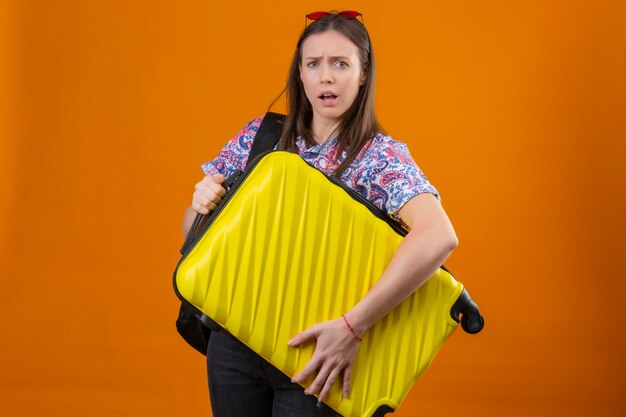 Foto gratuita mujer joven viajero con gafas de sol rojas en la cabeza con mochila con maleta mirando confundido sobre pared naranja