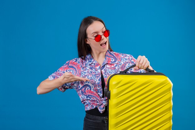 Mujer joven viajero con gafas de sol rojas con maleta amarilla apuntando con el brazo de la mano a int mirando alegre sobre la pared azul