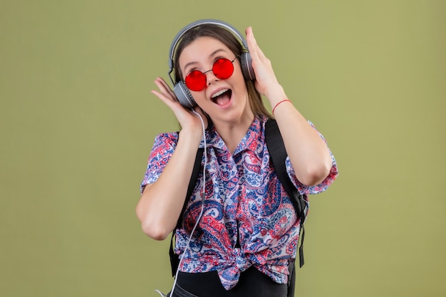 Mujer joven viajero con gafas de sol rojas y con mochila escuchando música con auriculares cantando con cara feliz de pie sobre fondo verde