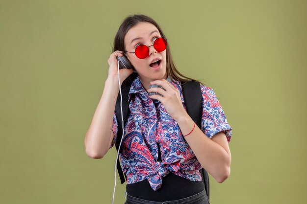 Mujer joven viajero con gafas de sol rojas y con mochila escuchando música con auriculares mirando sorprendido y feliz de pie sobre fondo verde