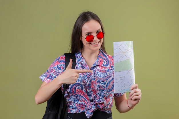 Mujer joven viajero con gafas de sol rojas y con mochila sosteniendo mapa apuntando con el dedo índice sonriendo alegremente sobre la pared azul