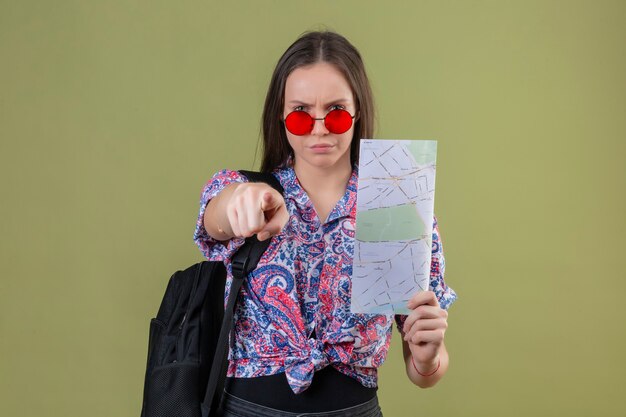 Mujer joven viajero con gafas de sol rojas y con mochila sosteniendo mapa disgustado apuntando a la cámara con el dedo con expresión enojada de pie sobre fondo verde