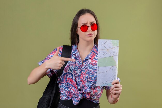 Mujer joven viajero con gafas de sol rojas y con mochila sosteniendo mapa disgustado apuntando hacia él con el dedo índice de pie sobre fondo verde