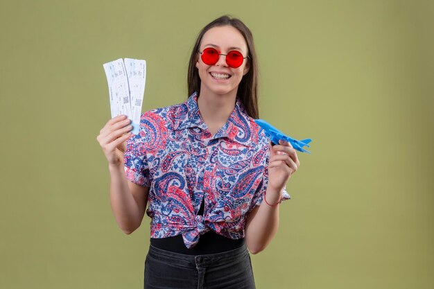 Mujer joven viajero con gafas de sol rojas sosteniendo boletos y avión de juguete mirando a un lado con cara feliz sonriendo alegremente de pie sobre fondo verde