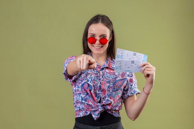 Mujer joven viajero con gafas de sol rojas sosteniendo boletos sonriendo con cara feliz apuntando con el dedo a la cámara de pie sobre fondo verde