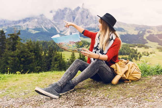 Mujer joven viajero con mochila y sombrero sentado en el césped y buscando la dirección correcta en el mapa