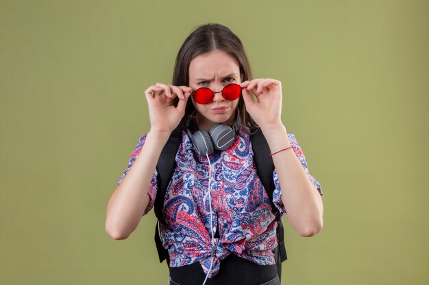 Foto gratuita mujer joven viajero de pie con mochila y auriculares con gafas de sol rojas tocándolos mirando a la cámara con expresión sospechosa de pie sobre fondo verde