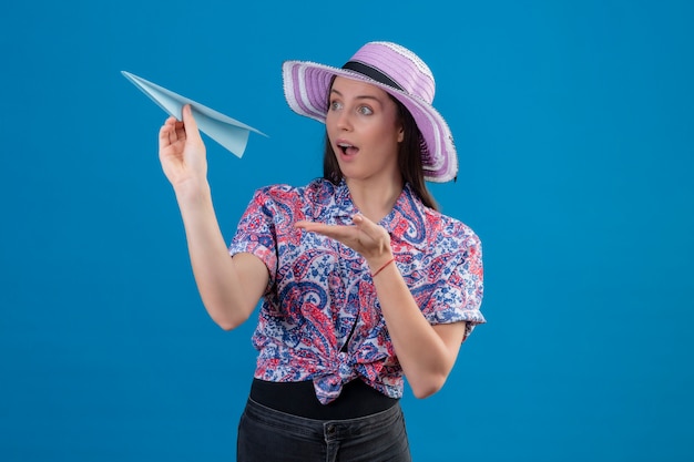 Mujer joven viajero en sombrero de verano con avión de papel apuntando con el brazo de la mano mirando sorprendido sobre la pared azul