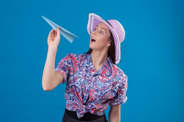 Mujer joven viajero en sombrero de verano con avión de papel juguetón y feliz sobre pared azul