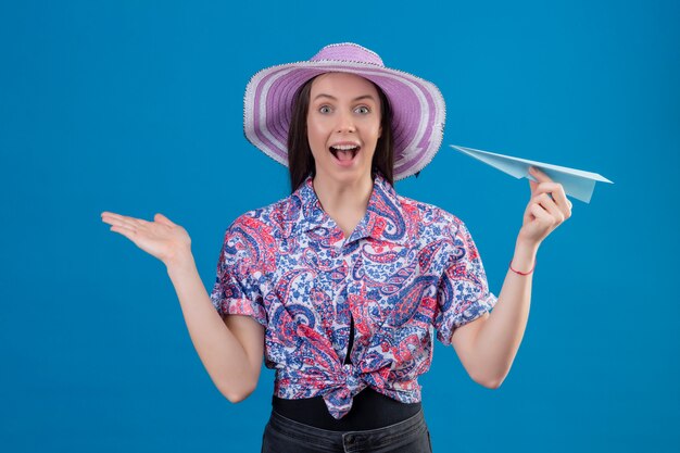 Mujer joven viajero en sombrero de verano con avión de papel mirando sorprendido y feliz con la mano levantada sobre la pared azul