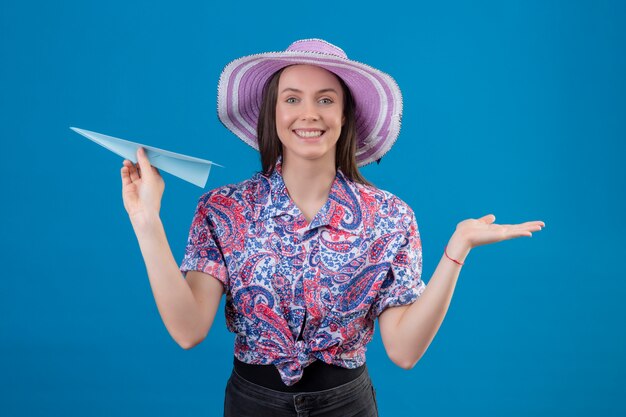 Mujer joven viajero en sombrero de verano con avión de papel que presenta con el brazo de la mano sonriendo alegremente con cara feliz sobre la pared azul