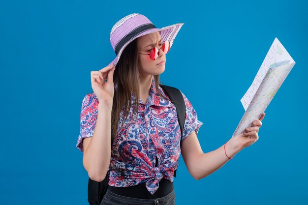 Mujer joven viajero en sombrero de verano con gafas de sol rojas con mapa mirando con cara de fruncir el ceño sobre la pared azul