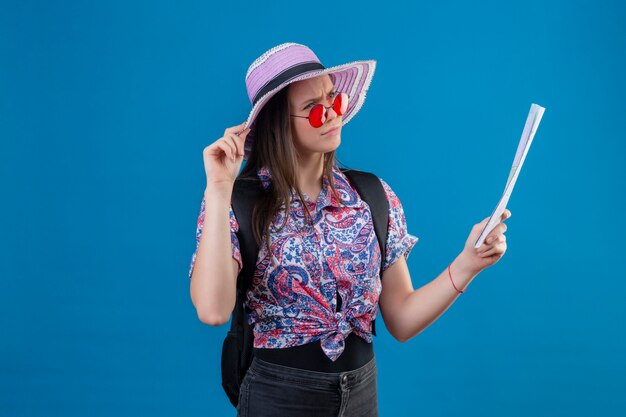 Mujer joven viajero con sombrero de verano con gafas de sol rojas sosteniendo el mapa mirando a un lado con expresión pensativa pensando tener dudas sobre fondo azul