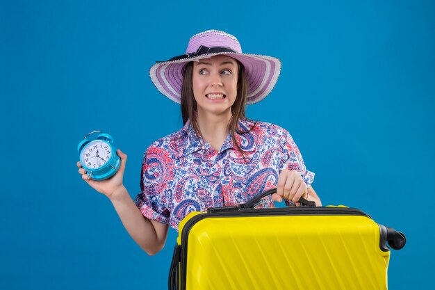 Mujer joven viajero con sombrero de verano de pie con maleta amarilla sosteniendo despertador estresado y nervioso sobre fondo azul.