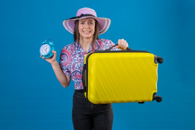 Mujer joven viajero con sombrero de verano de pie con maleta amarilla sosteniendo despertador estresado y nervioso sobre fondo azul.