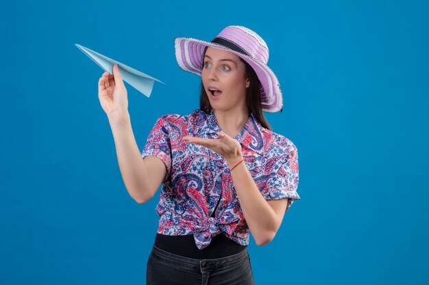 Mujer joven viajero con sombrero de verano sosteniendo avión de papel apuntando con el brazo de la mano hacia él mirando sorprendido de pie sobre fondo azul.