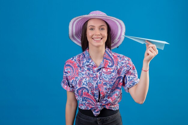 Mujer joven viajero con sombrero de verano sosteniendo avión de papel mirando a la cámara positiva y feliz sonriendo alegremente de pie sobre fondo azul.