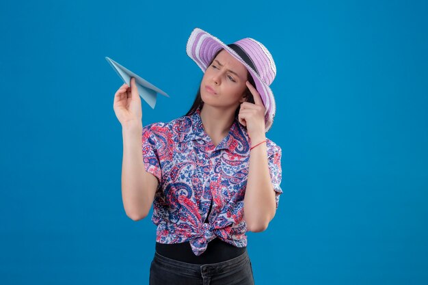 Mujer joven viajero con sombrero de verano sosteniendo avión de papel mirándolo con expresión pensativa teniendo dudas pensando de pie sobre fondo azul