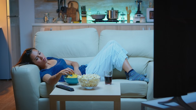 Mujer joven viendo la televisión y aburrida sentada en el sofá de la sala de estar en casa. Señora cansada, agotada y solitaria que se relaja viendo la televisión acostada en un cómodo sofá comiendo bocadillos a altas horas de la noche