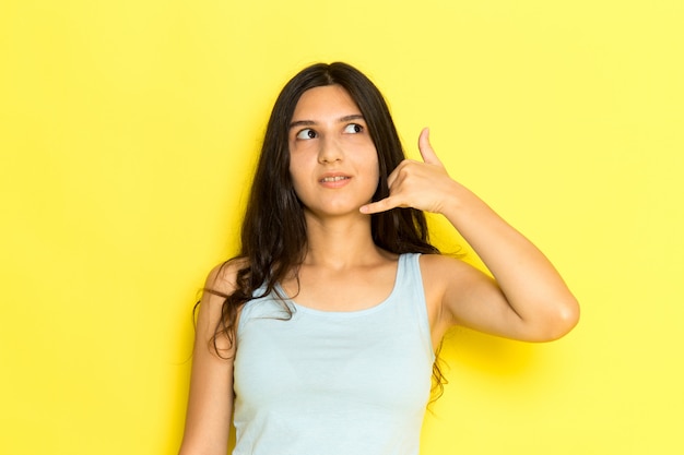 Una mujer joven de vista frontal en camisa azul posando y mostrando señal de llamada telefónica en el fondo amarillo pose de niña modelo belleza joven