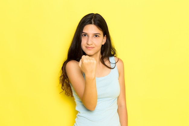 Una mujer joven de vista frontal en camisa azul posando y mostrando su puño sobre el fondo amarillo pose de niña modelo belleza joven