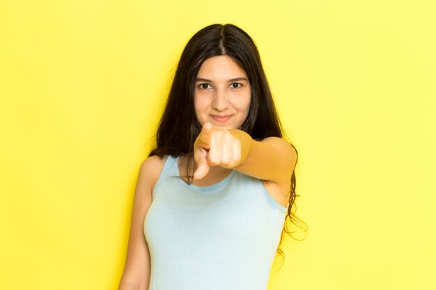 Una mujer joven de vista frontal en camisa azul posando y sonriendo con el dedo puntiagudo sobre el fondo amarillo pose de niña modelo belleza joven