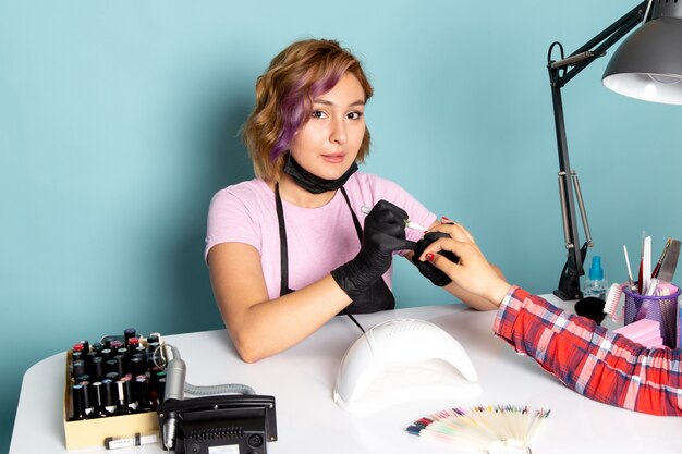 Una mujer joven de vista frontal manicura con guantes negros y máscara negra haciendo manicura en azul