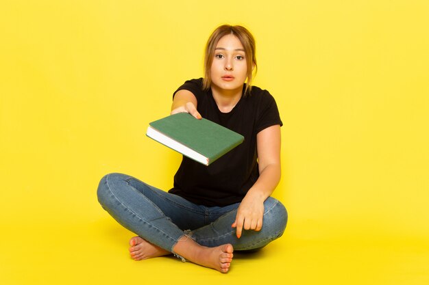 Una mujer joven de vista frontal sentada en camisa negra y jeans azul sosteniendo un cuaderno verde sobre amarillo