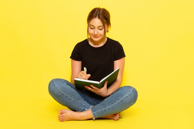 Una mujer joven de vista frontal sentada en camisa negra y jeans escribiendo notas con una sonrisa en su rostro en amarillo