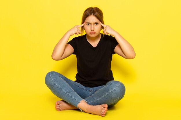 Una mujer joven de vista frontal sentada en camisa negra y jeans pensando tristemente en amarillo