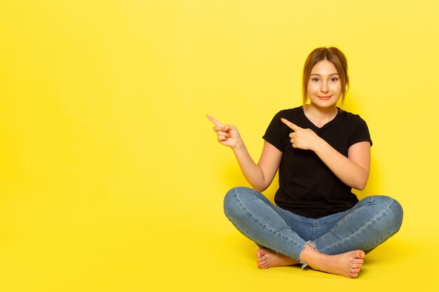 Una mujer joven de vista frontal sentada en camisa negra y jeans posando y señalando con una sonrisa en amarillo