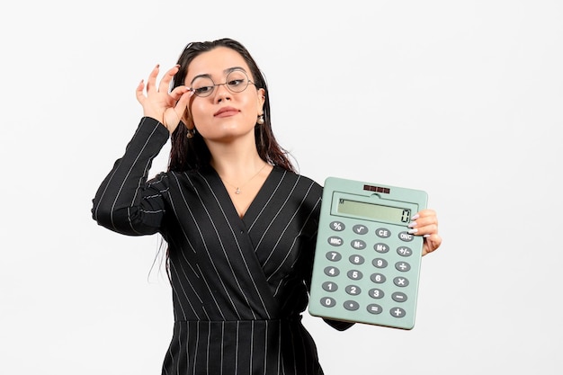 Mujer joven de vista frontal en traje oscuro estricto con gran calculadora en el trabajo de oficina de negocios de moda de mujer de belleza de piso blanco