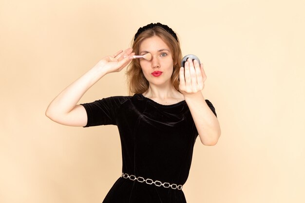 Una mujer joven de vista frontal en vestido negro con cinturón de cadena haciendo maquillaje sobre fondo beige maquillaje de rímel de moda