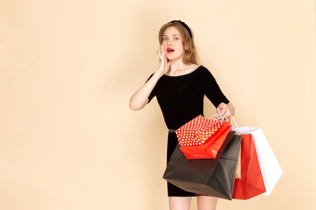 Una mujer joven de vista frontal en vestido negro con cinturón de cadena sosteniendo paquetes de compras en beige