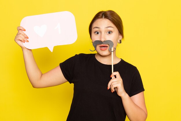 Una mujer joven de vista frontal en vestido negro posando con bigote falso con cartel blanco sobre amarillo