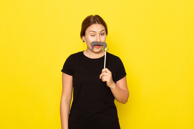 Una mujer joven de vista frontal en vestido negro posando con bigote falso con expresión divertida en amarillo