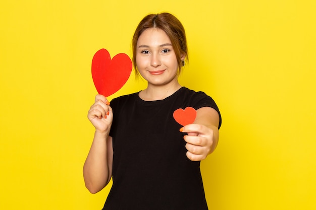 Una mujer joven de vista frontal en vestido negro posando con formas de corazón rojo sonriendo en amarillo
