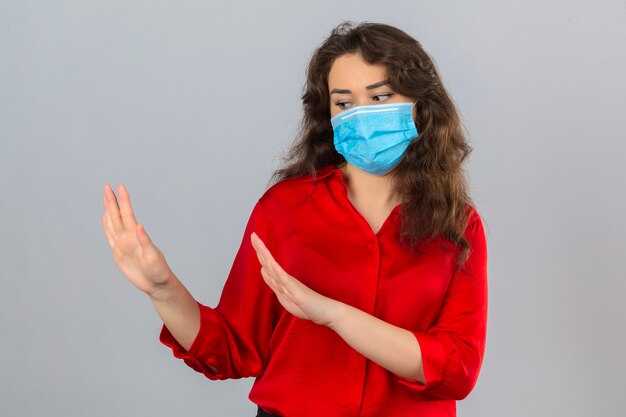 Mujer joven vistiendo blusa roja en máscara protectora médica sosteniendo las manos en alto diciendo que no se acerquen sobre fondo blanco aislado