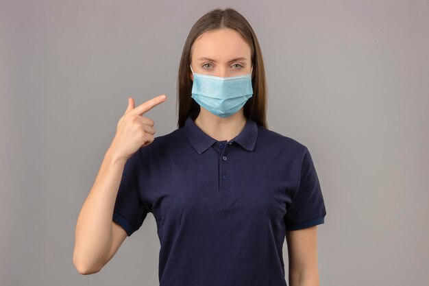 Mujer joven vistiendo camisa polo azul en máscara médica protectora apuntando con el dedo en su máscara con cara seria mirando a la cámara de pie sobre fondo gris claro