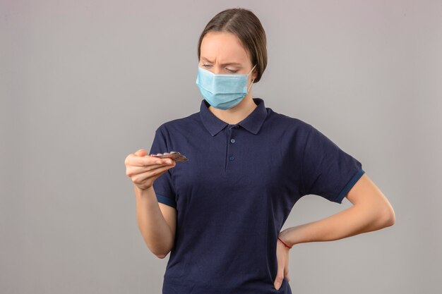 Mujer joven vistiendo camisa polo azul en máscara médica protectora con pastillas de ampolla en la mano mirando las píldoras con cara seria de pie sobre fondo gris claro