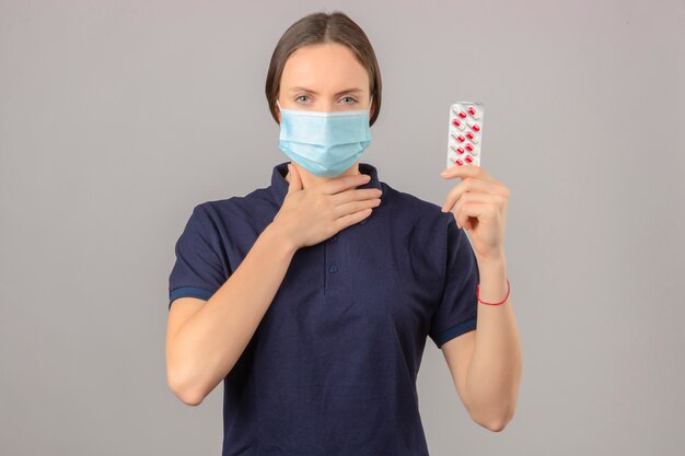 Mujer joven vistiendo una camisa polo azul en máscara médica protectora con pastillas de ampolla en la mano tocando su cuello de pie sobre fondo gris claro