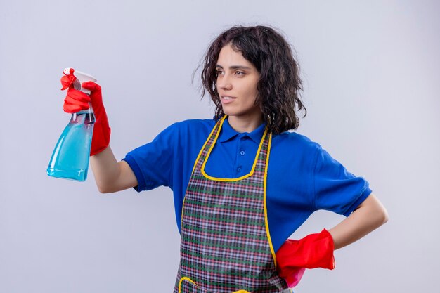 Mujer joven vistiendo delantal y guantes de goma de pie con spray limpiador listo para limpiar sobre fondo blanco aislado