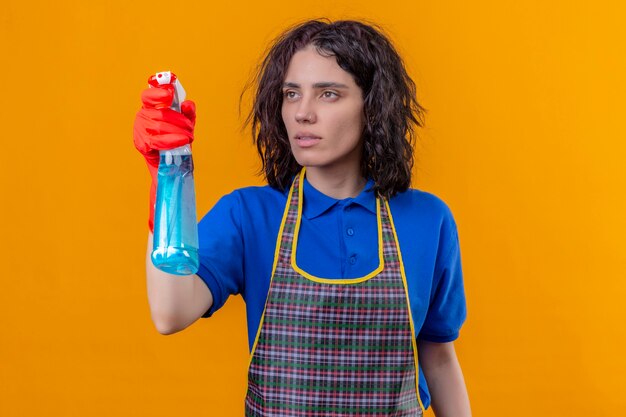 Mujer joven vistiendo delantal y guantes de goma sosteniendo spray de limpieza mirando a un lado con cara seria listo para limpiar de pie sobre fondo naranja